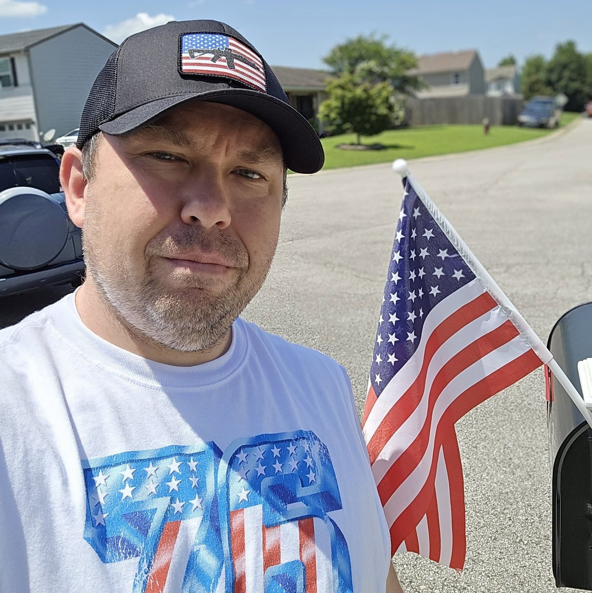 Man in a patriotic hat and shirt stands outside next to a small American flag.